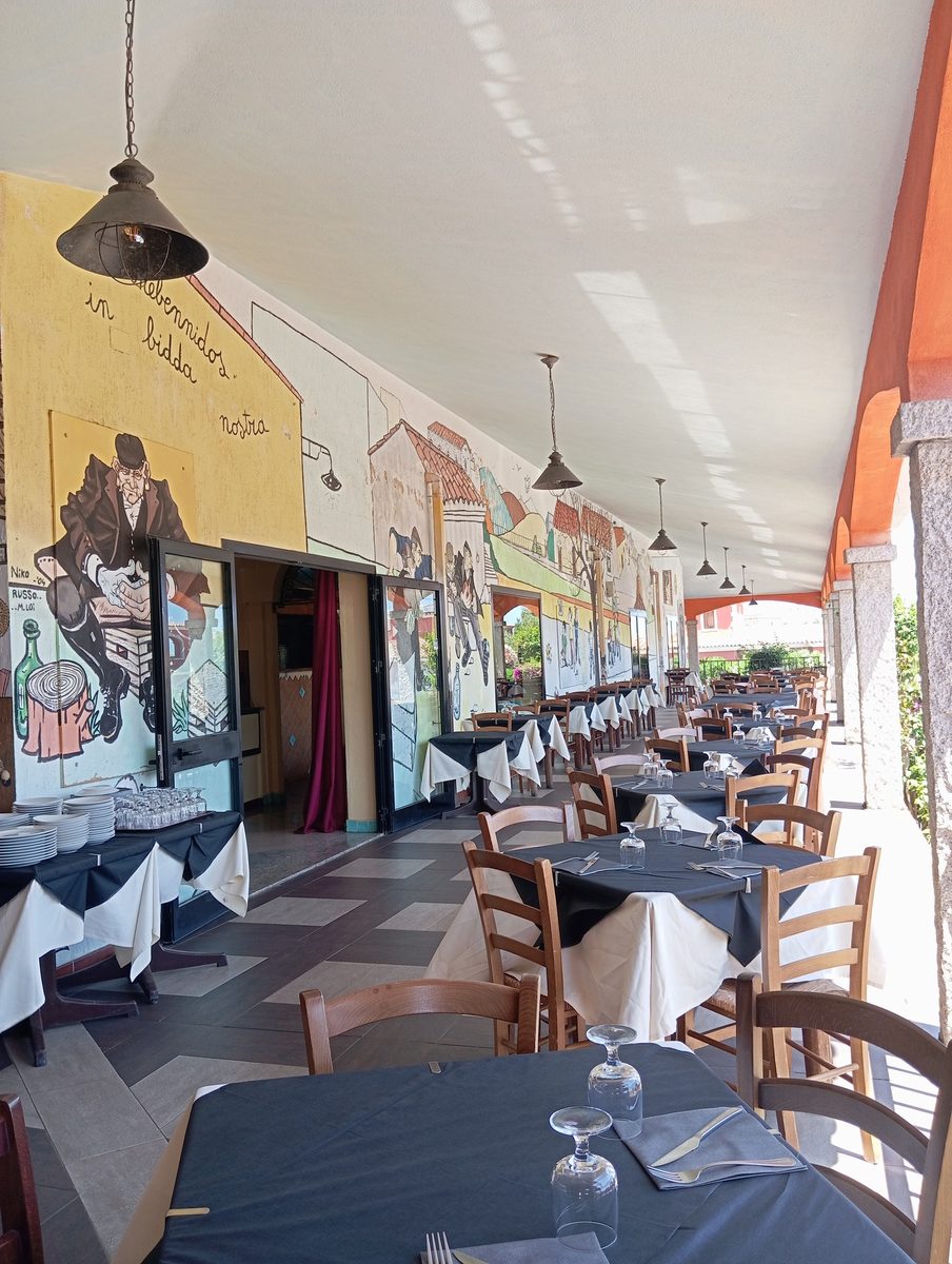 Dehor estivo del Ristorante Pizzeria La Mesenda a San Teodoro, Sardegna – tavoli apparecchiati con murales sardi sullo sfondo